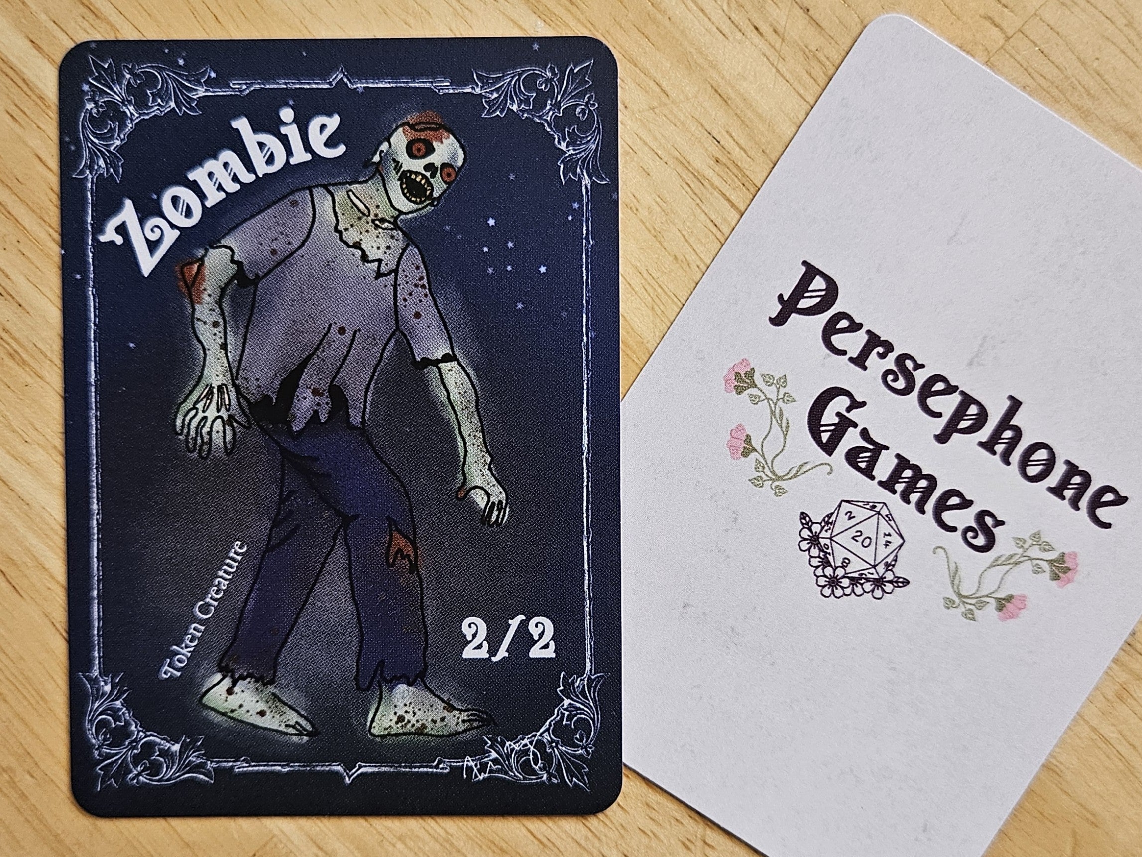 Illustrated 2/2 zombie token card featuring a bloodied, hobbling zombie glowing red eyes, torn dark blue pants and gray t-shirt that is falling apart.